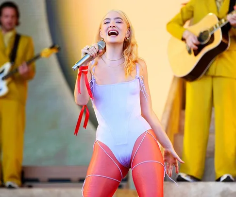 Singer performing on stage with blonde hair, wearing a white outfit, holding a microphone, musicians in background.