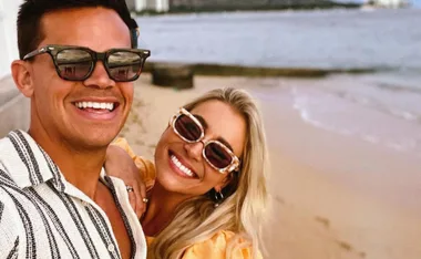 Couple smiling on a beach, wearing sunglasses, with ocean and buildings in the background.