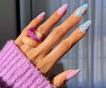 A hand with colorful manicured nails in purple and blue patterns, wearing a chunky ring, and a purple sweater.