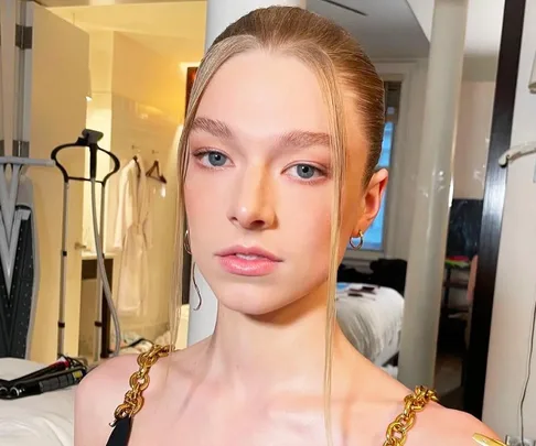 Person with light hair tied back, wearing gold chain-strap top, in a room with an ironing board and laundry.