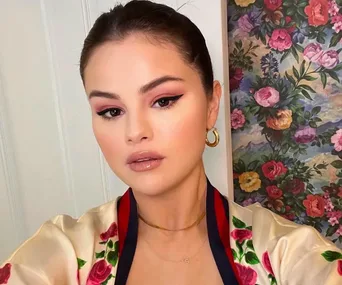 A woman with makeup in front of a floral wallpaper, wearing a floral robe and gold earrings.