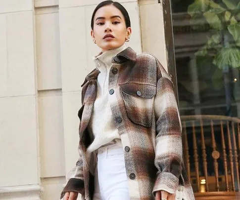 Woman in plaid shirt jacket over turtleneck sweater, standing outdoors against a building.