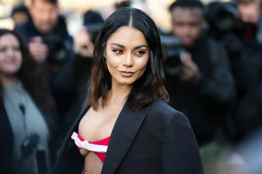 Vanessa Hudgens at a fashion event, wearing a black blazer over a red and white top, while photographers capture her.