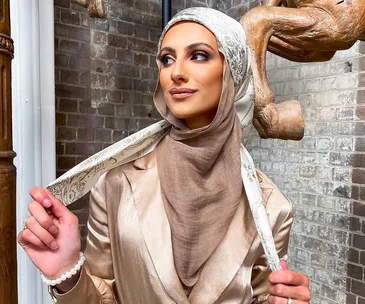 Woman in a stylish beige hijab and satin outfit with a printed scarf, against a rustic brick background.