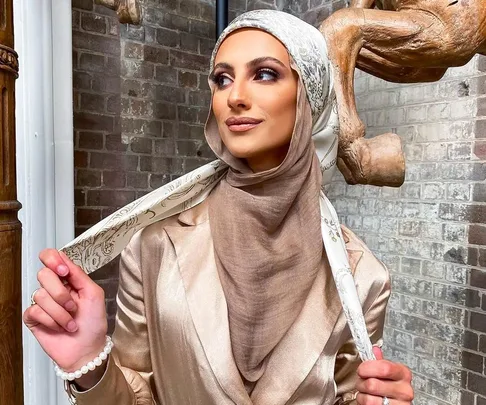 Woman in a stylish beige hijab and satin outfit with a printed scarf, against a rustic brick background.