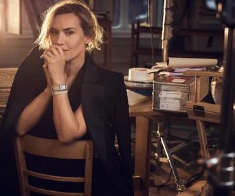 Person in black blazer with short hair, wearing a watch, sits on a wooden chair in a warmly lit room.
