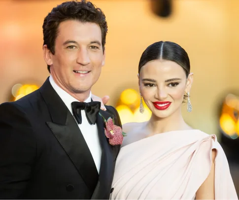 A man in a tuxedo with a flower on the lapel and a woman in a white gown with red lipstick pose at a formal event.