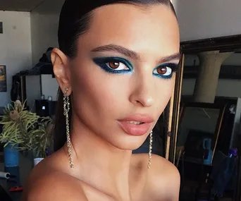 Woman wearing bold blue eyeshadow and long earrings in a well-lit room.