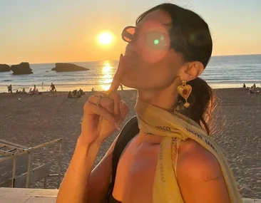 Person with sunglasses and scarf poses at sunset beach, kissing gesture, wearing heart-shaped earrings.