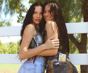 Two women smiling and hugging outdoors, wearing casual outfits with phones in their back pockets.