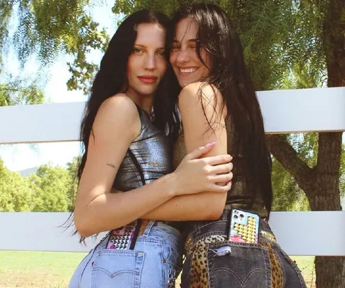 Two women smiling and hugging outdoors, wearing casual outfits with phones in their back pockets.