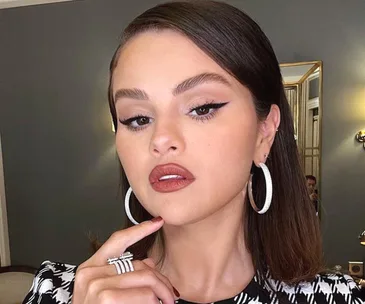 Woman with hoop earrings and makeup poses; dark slicked hair, patterned top, in a room with warm lighting.