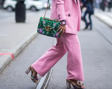 Person in a pink suit walks, carrying a green floral Gucci bag, wearing colorful high heels on a city street.