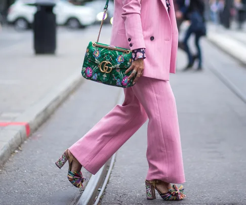 Person in a pink suit walks, carrying a green floral Gucci bag, wearing colorful high heels on a city street.