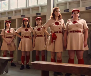 A group of women in vintage baseball uniforms with red caps stands in a locker room.