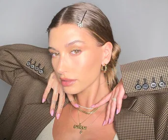 Model with slicked-back hair wears a plaid blazer, showcasing pink nails and layered gold necklaces.