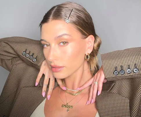 Model with slicked-back hair wears a plaid blazer, showcasing pink nails and layered gold necklaces.