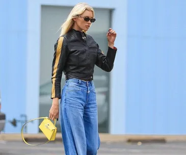 A person in a black jacket and wide-leg jeans walks with a yellow handbag, wearing sunglasses outdoors.