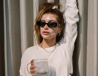 A woman with sunglasses and face cream holds a glass with ice, posing against a curtain backdrop.