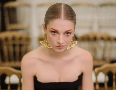 A person with slicked-back hair wearing large gold earrings and a strapless top, sitting on a chair in a fashion show setting.