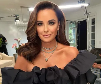 Woman with long brown hair and layered necklaces in a glamorous black outfit, smiling indoors with lights and flowers behind.