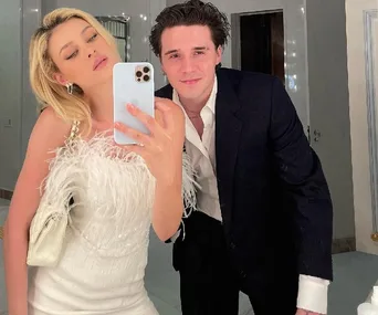 A couple posing together, the woman in a white feathered dress holding a phone, the man in a dark suit smiling in a mirror selfie.