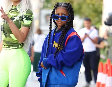 A young girl in braided hair wears oversized blue sunglasses and a blue jacket at a street fashion event.