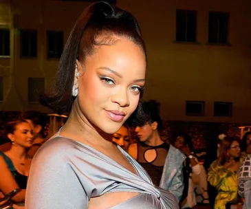 Rihanna in a stylish outfit at an event, surrounded by attendees, with a confident smile and hair in a sleek updo.