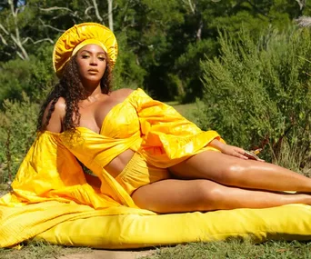 A woman lounges outdoors on a yellow cushion, wearing a bright yellow outfit and matching headpiece, surrounded by greenery.