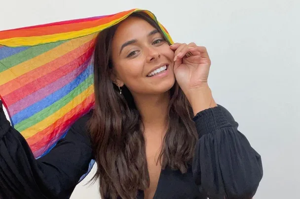 Woman smiling, holding a rainbow fabric over her head against a plain background.