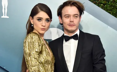 A woman in a gold dress and a man in a tuxedo pose together on a red carpet event backdrop.