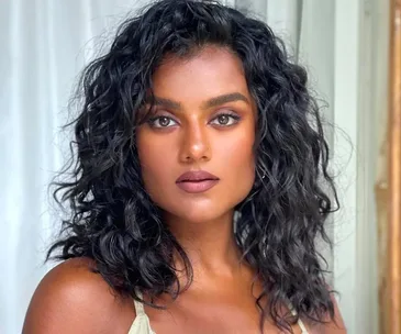 A woman with curly dark hair and natural makeup gazes at the camera against a soft background.