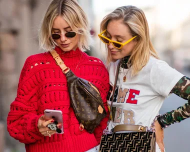 Two women in trendy outfits and sunglasses look at a phone together. One wears a red sweater and the other a layered shirt.