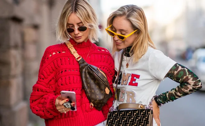 Two women in trendy outfits and sunglasses look at a phone together. One wears a red sweater and the other a layered shirt.