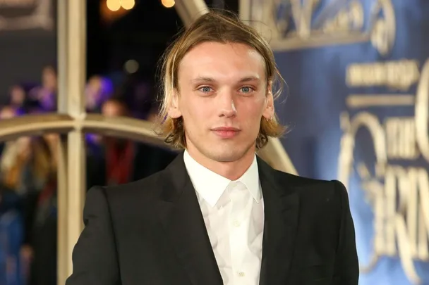 A man in a suit with long hair at a formal event, blue backdrop with blurred writing.