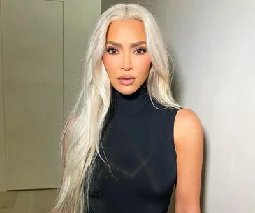 A woman with long platinum blonde hair wearing a black sleeveless turtleneck top stands indoors against a plain wall.