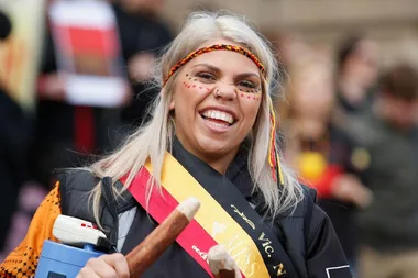 Smiling woman with face paint and traditional headband at NAIDOC Week event, holding boomerangs.