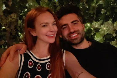 A smiling couple sitting closely with an arm around each other, surrounded by greenery in the background.