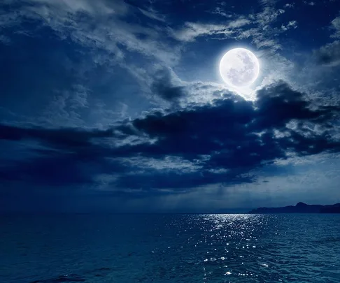 Full moon illuminating a calm night ocean, partially covered by clouds.