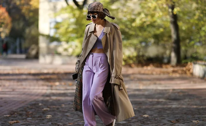 Person walking outdoors in a beige trench coat, purple top, lilac pants, headscarf, and sunglasses.