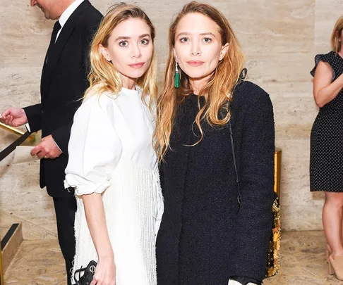 Two women with long hair, one in a white dress and the other in a black coat, pose in a formal setting.