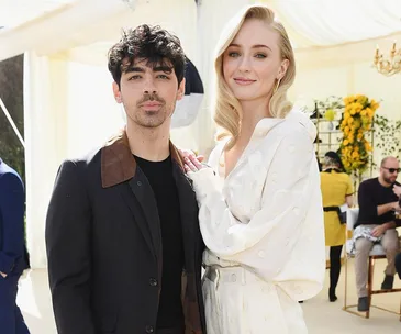 A couple standing together at a sunny outdoor event, both smiling and dressed elegantly.