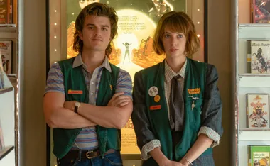 Steve and Robin from "Stranger Things" wearing Family Video uniforms, standing in front of a movie poster display.