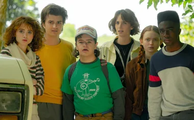 Six "Stranger Things" characters standing together outdoors, looking concerned, beside a vehicle.