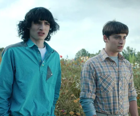 Two teenage boys stand in a field, one with curly black hair in a teal shirt, the other with short brown hair in a plaid shirt.