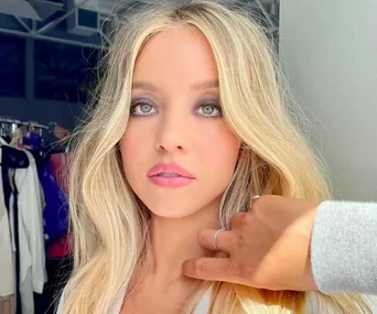 Woman with long blonde hair getting makeup touched up, wearing a light top, standing indoors with clothing racks in background.