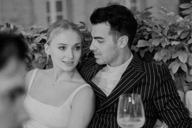 A couple sits together on a bench, gazing at each other affectionately, surrounded by foliage.