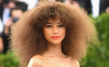 A woman with voluminous curly brown hair and orange lip color poses outdoors in formal attire.