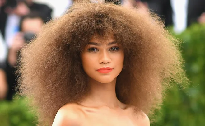 A woman with voluminous curly brown hair and orange lip color poses outdoors in formal attire.