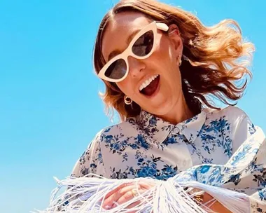 Woman in white-framed sunglasses and floral shirt smiling under a bright blue sky.